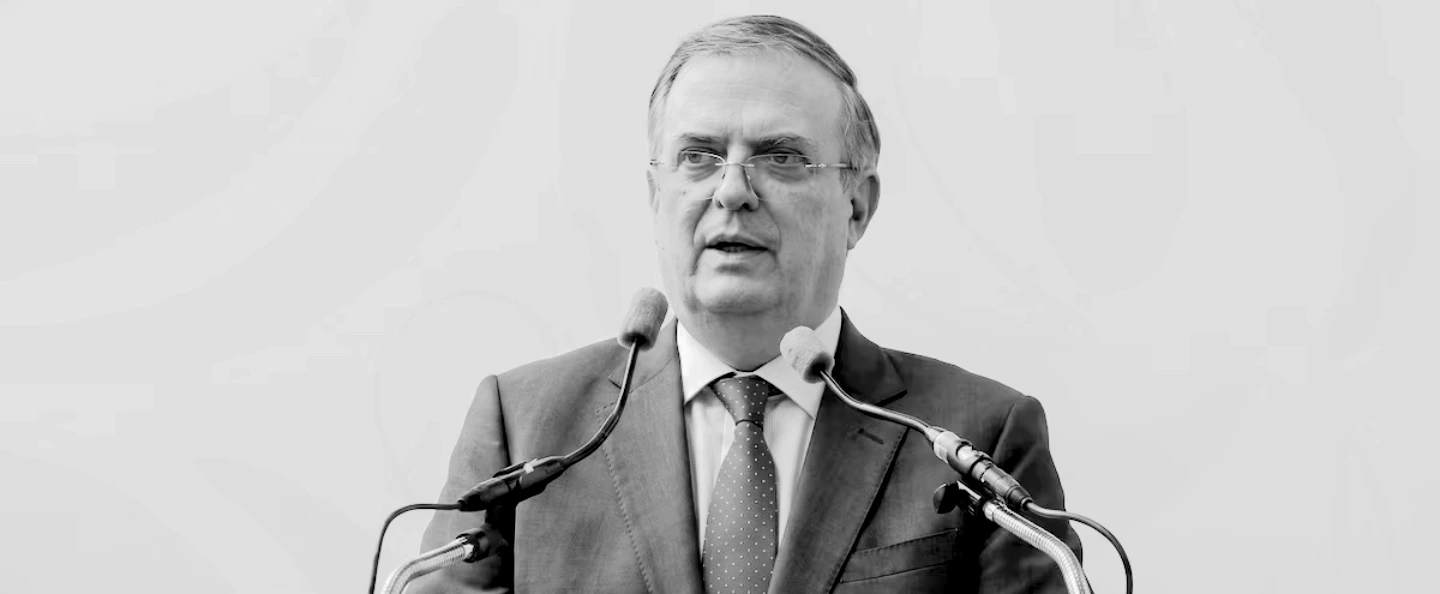 Marcelo Ebrard habla ante un micrófono en un evento empresarial, con un fondo que muestra productos de exportación