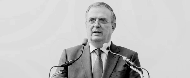 Marcelo Ebrard habla ante un micrófono en un evento empresarial, con un fondo que muestra productos de exportación